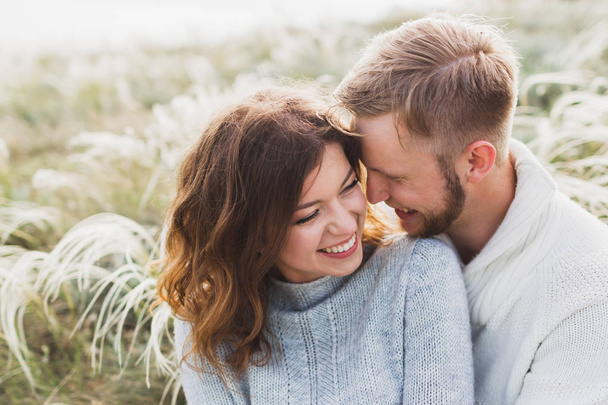 Счастливая молодая любящая пара, сидящая на лугу из перьев, смеющаяся и обнимающаяся, свитер и джинсы повседневного стиля
 - Фото, изображение