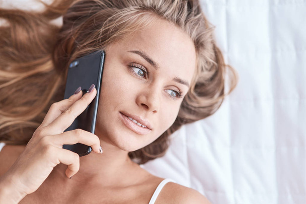 Thoughtful glance. Nice attractive young woman lying in bed and talking on phone while looking aside - Foto, imagen