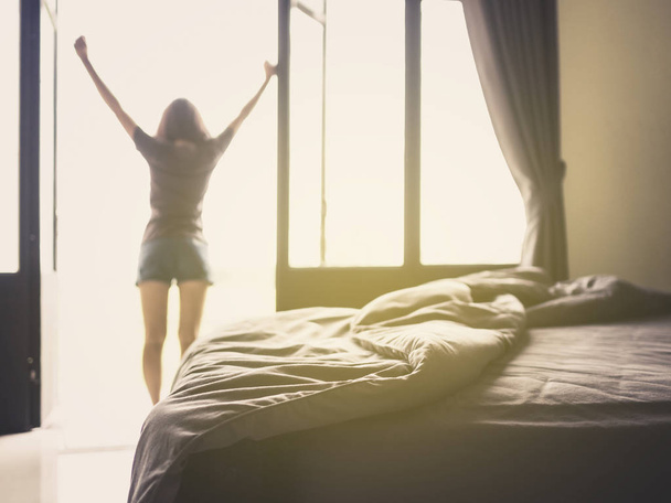 Путаница одеяло и кровать в роскошной спальне и здоровой молодой женщины растяжения и отдыха у окна после пробуждения утром праздника. Селективный фокус
. - Фото, изображение