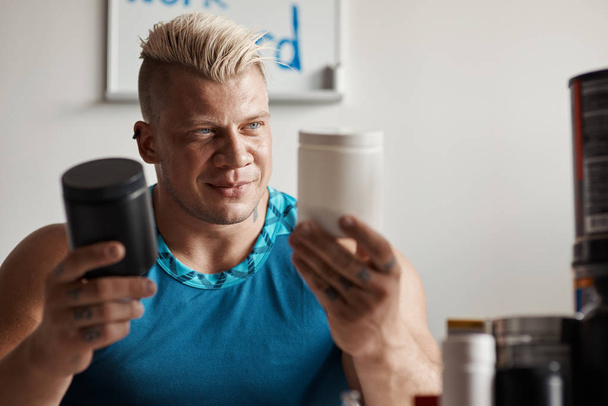 What to choose. Waist up of a professional serious strongman holding supplements and sitting at the table while making a choice - Valokuva, kuva