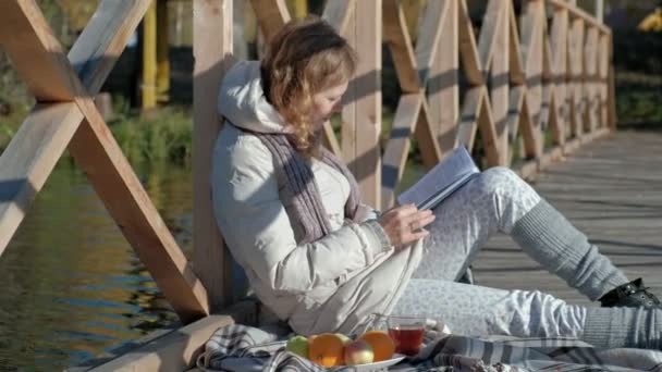 una mujer en el puente cerca del río de otoño lee un libro y bebe té
 - Imágenes, Vídeo