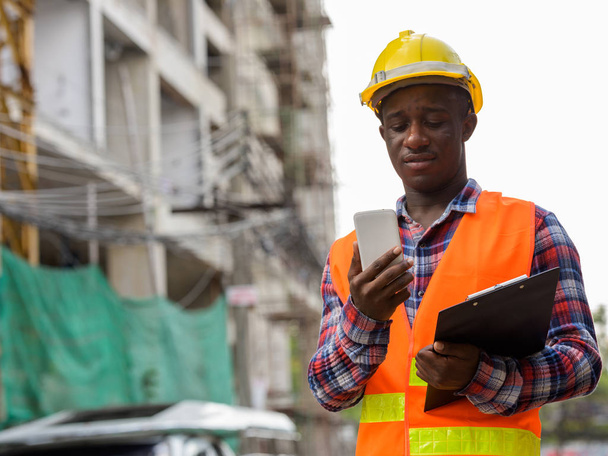 Jonge zwarte Afrikaanse man bouwvakker met klembord tijdens het gebruik van mobiele telefoon op de bouwplaats - Foto, afbeelding