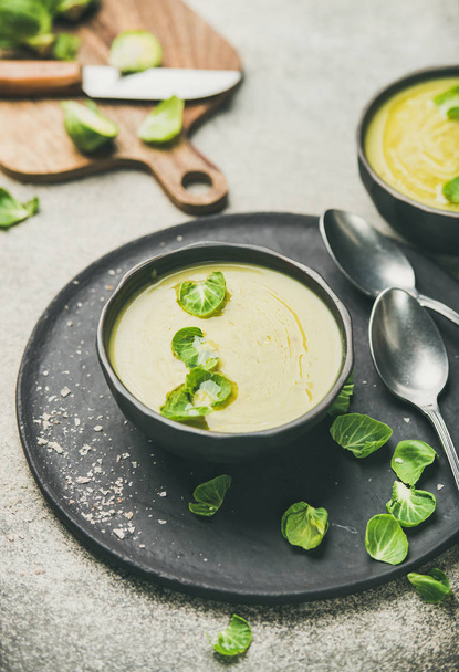 Seasonal brussels sprouts vegetable cream soup in dark bowl and fresh green brussel sprouts over grey concrete background, vertical composition. Vegan, vegetarian, healthy, dieting food concept - Photo, image