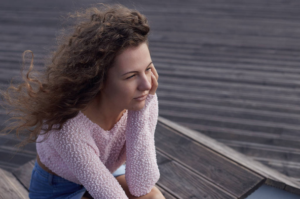 Authentic portrait of young Caucasian attractive female, watching  sunset, looking afar. Wind is messing long curly hair. Close up shot of thoughtful woman dressed in pink top and blue denim skirt. - Фото, изображение