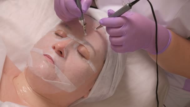 kosmetisches Verfahren zur Gesichtsmassage mit einem elektrischen Gerät. Eine Frau mit weißer Maske befindet sich in einer Arztpraxis. die Hände einer Kosmetikerin in Handschuhen glätten mit Hilfe von zwei Elektroden Falten. - Filmmaterial, Video