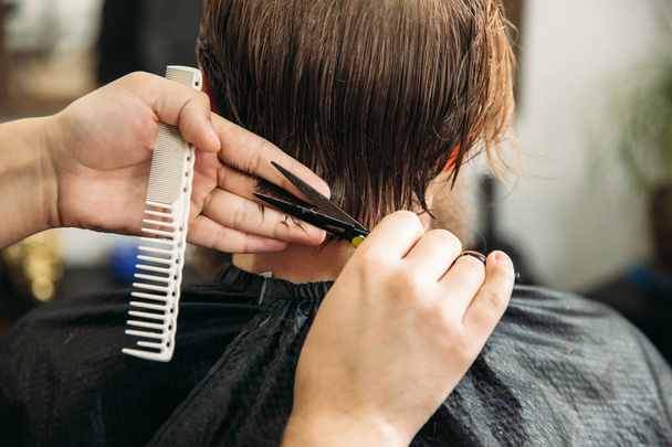 Руки молодого парикмахера, стригущего волосы привлекательному бородатому мужчине в парикмахерской
 - Фото, изображение
