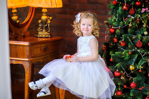 beautiful little girl in a dress sits in an armchair with a gift in hand, near the holiday tree - 写真・画像