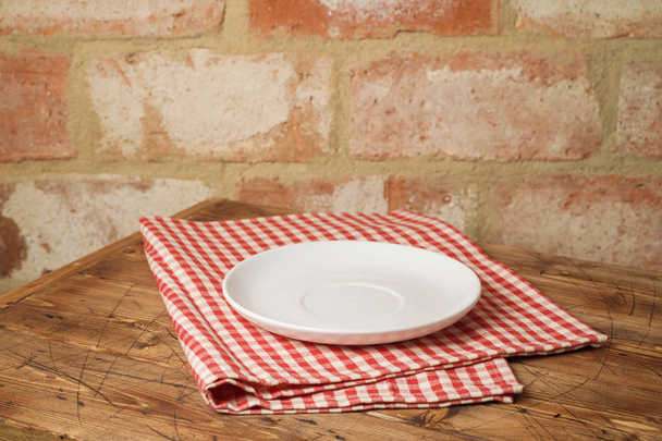 Empty white plate on wooden table with tablecloth over brick wall background - Fotó, kép