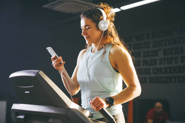 Theme sport and music. A beautiful Caucasian woman running in gym on treadmill. On head big white headphones, the girl listens to music during a cardio workout for weight loss and uses phone in hand. - Photo, Image