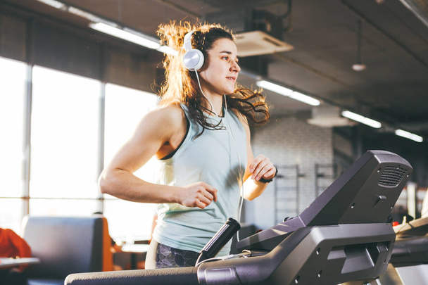 Theme is sport and music. A beautiful inflated woman runs in the gym on a treadmill. On her head are big white headphones, the girl listens to music during a cardio workout for weight loss. - Φωτογραφία, εικόνα