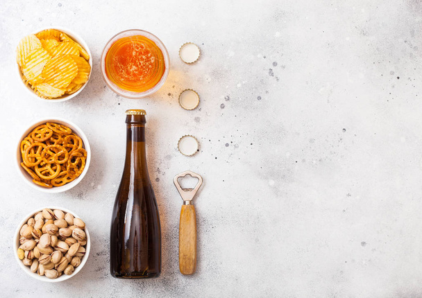 Glass and bottle of craft lager beer with snack on stone kitchen background. Pretzel and crisps and pistachio in white ceramic bowl - Фото, зображення