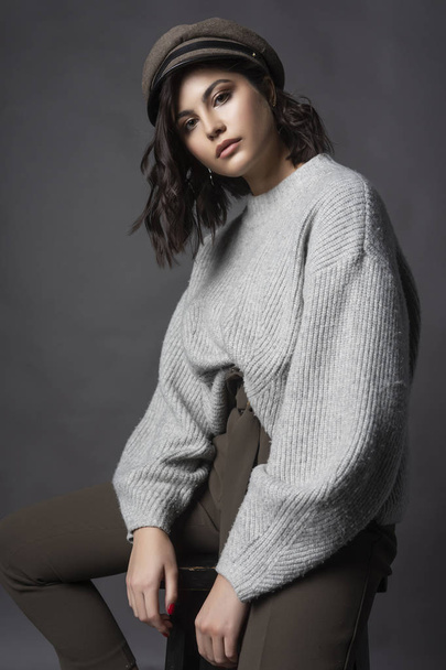 Beautiful brunette girl wearing casual style knitted sweater, brown pants and a hat, sits on a stool on a gray background. Natural nude makeup. Fashion, advertising and commercial design. Copy space. - Φωτογραφία, εικόνα