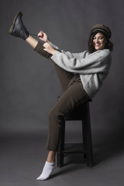 Beautiful smiling brunette girl wearing a casual style sweater, pants and cap puts a shoe on her leg on a gray background. Natural nude makeup. Fashion, advertising and commercial design. Copy space. - Photo, Image