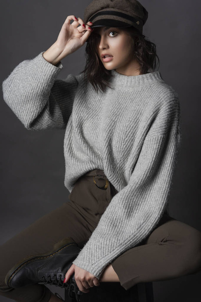 Beautiful surprised brunette girl wearing casual style sweater, pants, cap and a boots sits with her leg up on a stool and holds her hand over the visor on a gray background. Nude makeup. Copy space - 写真・画像
