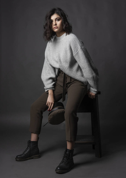 Beautiful brunette girl wearing a casual style sweater, pants, cap and a boots sits on a stool and holds a cap in her hand on a gray background. Nude natural makeup Fashion design. Copy space - Фото, изображение
