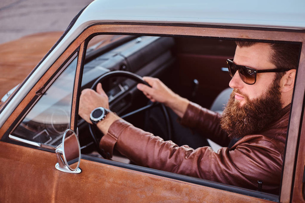 Bearded male in sunglasses dressed in brown leather jacket driving a retro car. - 写真・画像