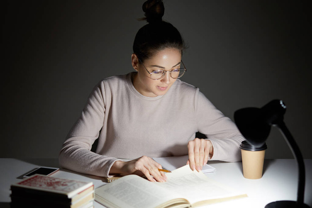 Shot of serious young Caucasian woman has focused look into textbook, finds necessary information, holds pencil, wears round big spectacles, sits in darkness, dressed casually. Education concept - Foto, Imagen