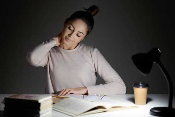 Fatigue female has pain in neck after working long time at desktop, wants to have rest, prepares for entering unversity, learns material from scientific literature, poses in dark room alone. - Foto, afbeelding