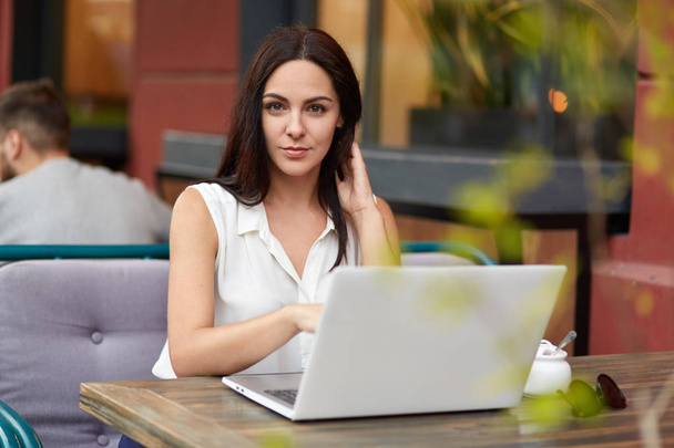Outdoor shot of beautiful brunette woman in white blouse watches educational webinar to get more knowledge, sits in front of opened laptop in cafeteria, has e learning, connected to free internet - Foto, Imagem