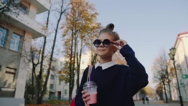 Pretty hipster teen with red bag drinks milkshake from a plastic cup walking street between buildings. Cute girl in sunglasses drinks a beverage through a straw. - Video, Çekim
