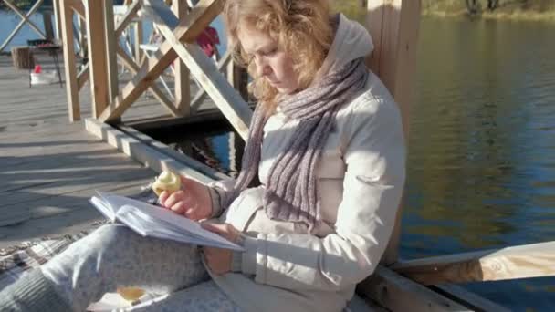 mujer en el puente cerca del río de otoño lee un libro y se come una manzana
 - Imágenes, Vídeo