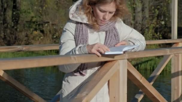 woman on the bridge near the autumn river reads a book - Кадри, відео