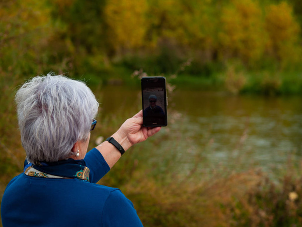 Ένας ανώτερος γυναίκα με γκρίζα μαλλιά βγάζει selfies φωτογραφίες στην όχθη το φθινόπωρο - Φωτογραφία, εικόνα