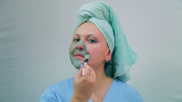 A young woman with a towel on her head applies a mud mask on her face with a brush. Looking at the camera. - Séquence, vidéo