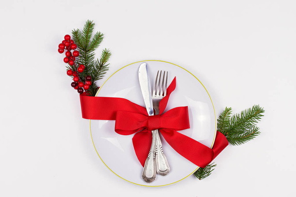 Karácsonyi összetétele, tányér, evőeszköz, fenyő ágak, szalag, piros bogyós gyümölcsök, a fehér asztal. Téli ünnepek és ünnepi háttér. Karácsonyi vacsora, szilveszteri étel ebéd. szemközti nézet - Fotó, kép
