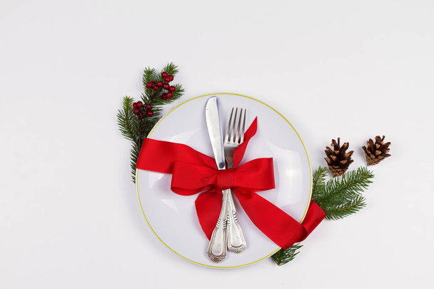 Christmas composition with plate, cutlery, pine branches, ribbon and red berries on white table. Winter holidays and festive background. Christmas eve dinner, New Year food lunch.  top view - Fotó, kép