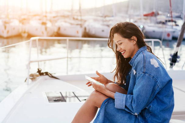 Young beautiful girl is sitting on the deck of a yacht with a smartphone - Photo, Image
