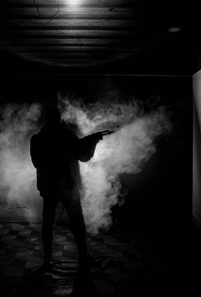 アサルトライフルの暗いトーンの霧の背景または危険な山賊の手に銃を保持している攻撃する準備を持つ男のシルエット。武器のテーマの装飾が施されたテロリストを撮影 - 写真・画像