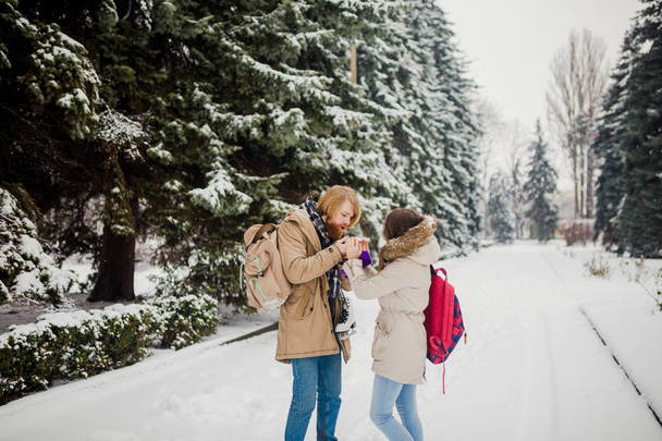 Date young couple in winter. Young Caucasian man with beard and long hair holding in hands the hands of girl with knitted gloves and warming and kissing them in snow park in winter is Valentines Day. - 写真・画像
