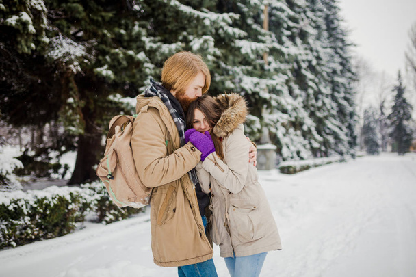 Νεαρό ζευγάρι ημερομηνία το χειμώνα. Καυκάσιος νέος άνθρωπος με γένια και μακριά μαλλιά, κρατώντας στα χέρια τα χέρια του κοριτσιού με πλεκτά γάντια και υπερθέρμανση του πλανήτη και τους φιλιά στο πάρκο χιονιού το χειμώνα είναι ημέρα του Αγίου Βαλεντίνου. - Φωτογραφία, εικόνα