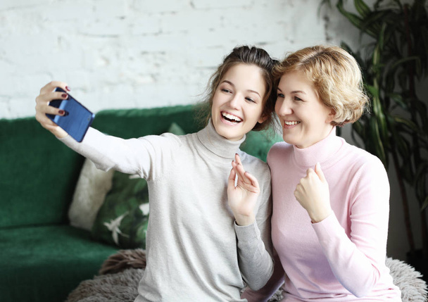 Portrait of beautiful mature mother and her daughter making a selfie using smart phone and smiling, home and happy. - Foto, afbeelding