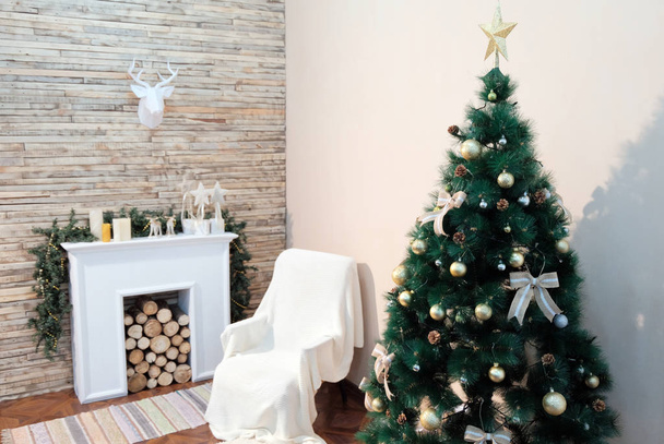 Christmas tree stands near the bedside table and fireplace against the white wall. Paper deer head hanging on the wall of wooden planks. Theme happy new year. - Photo, Image