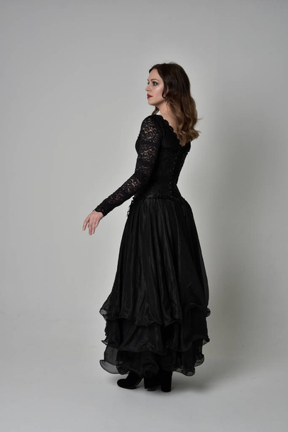 full length portrait of brunette girl wearing long black lace gown wit corset. standing pose with back to the camera. isolated on grey studio background. - Fotó, kép