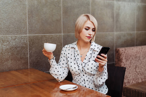Young business woman in cafe drinking coffee and looking at smartphone - Фото, изображение