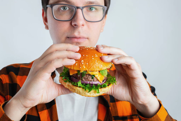 A young guy with glasses holding a fresh Burger. A very hungry student eats fast food. Hot helpful food. The concept of gluttony and unhealthy diet. With copy space for text. - Фото, изображение