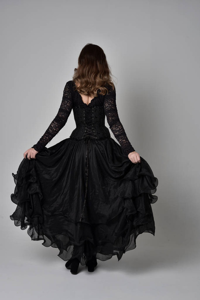 full length portrait of brunette girl wearing long black lace gown wit corset. standing pose with back to the camera. isolated on grey studio background. - Fotografie, Obrázek