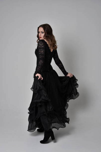 full length portrait of brunette girl wearing long black lace gown wit corset. standing pose with back to the camera. isolated on grey studio background. - Photo, Image