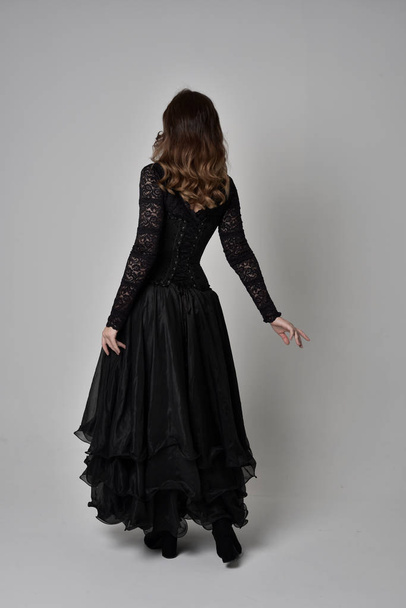 full length portrait of brunette girl wearing long black lace gown wit corset. standing pose with back to the camera. isolated on grey studio background. - Fotó, kép