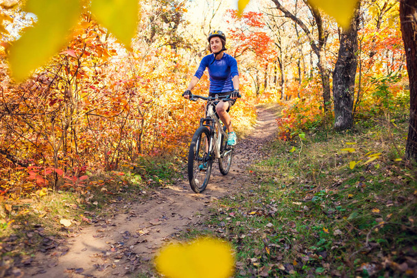 The girl with a backpack rides a bike in the autumn park. Slender woman trains in nature. Sports in the forest. Tourist rides on a dirt trail. Traveling by bike. - Photo, Image