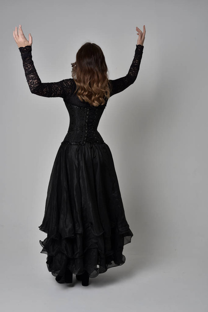 full length portrait of brunette girl wearing long black lace gown wit corset. standing pose with back to the camera. isolated on grey studio background. - Foto, Imagem