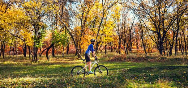 The girl with a backpack rides a bike in the autumn park. Slender woman trains in nature. Sports in the forest. Tourist rides on a dirt trail. Traveling by bike. - Photo, Image