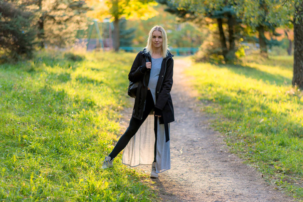 Photo portrait of a beautiful blonde girl in the park on the street in the fall in the sunset sun evening. She is standing in front of the camera, smiling and looking happy. - Foto, Imagen