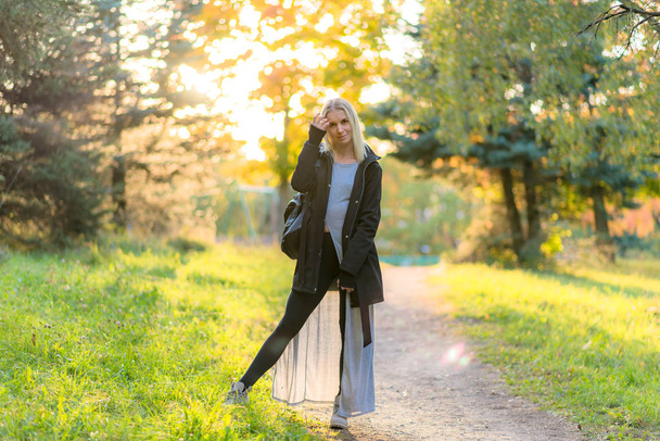 Photo portrait of a beautiful blonde girl in the park on the street in the fall in the sunset sun evening. She is standing in front of the camera, smiling and looking happy. - Foto, afbeelding