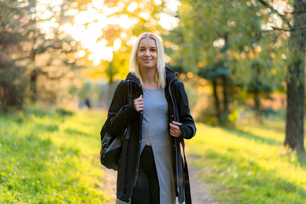 Photo portrait of a beautiful blonde girl in the park on the street in the fall in the sunset sun evening. She is standing in front of the camera, smiling and looking happy. - 写真・画像