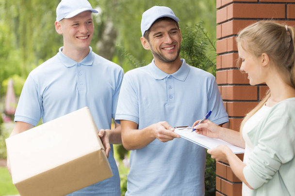 Two handsome couriers in blue uniforms delivering a parcel to a young pretty woman - Photo, Image