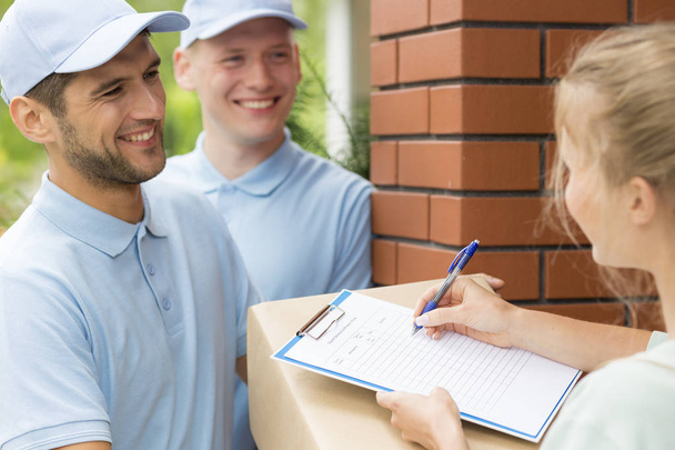 Smiling friendly couriers in blue uniforms and woman signing receipt of package delivery - Фото, изображение